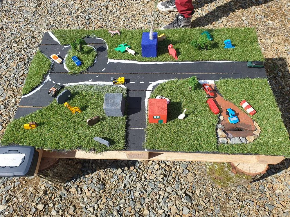 Ein selbst gebauter Spieltisch aus einer Palette: eine Miniatur-Straßenlandschaft mit Kunstrasen, aufgemalten Straßen, kleinen Häusern, Bäumen, Tieren und Spielzeugautos.
