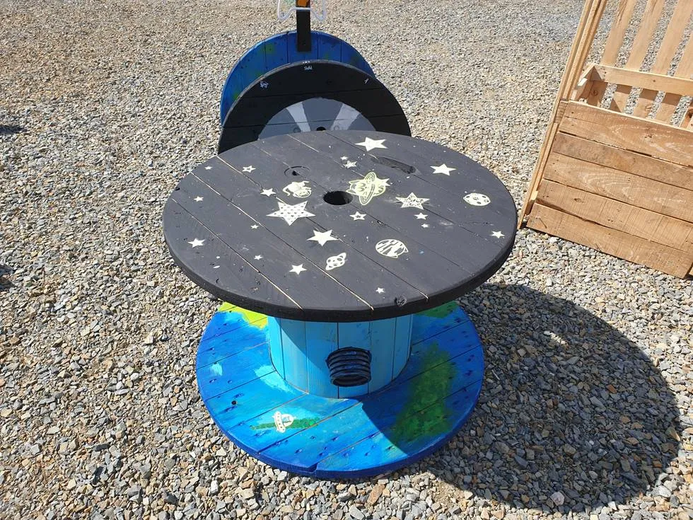 Ein Spieltisch aus einer Kabeltrommel im Weltraum-Look mit aufgemalten Sternen und Planeten.